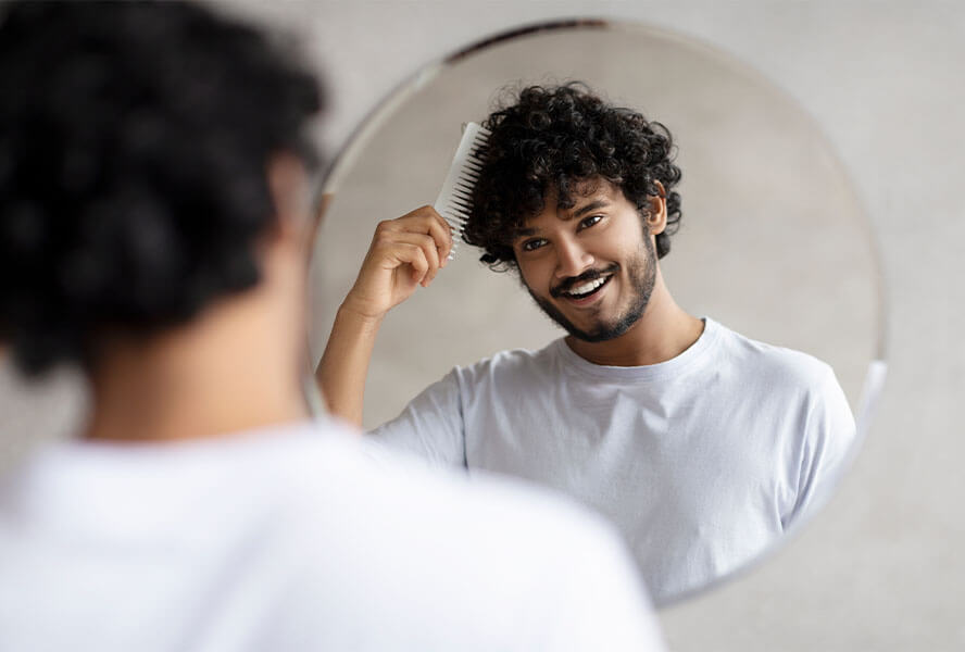 Mann mit dunklen Locken steht vor dem Spiegel und kämmt seine Haare mit einem Kamm