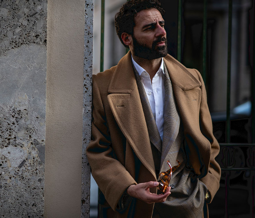 Dunkelhaariger Mann mit Sonnenbrille, rosafarbenem Hemd und beiger Chino lehnt an einer Wand